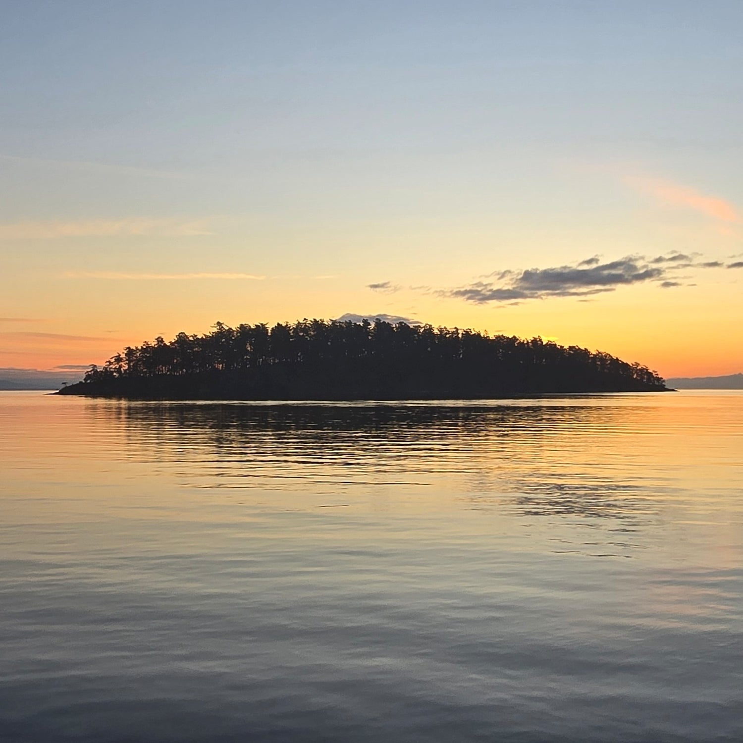 A tranquil sunset seascape with a small tree-covered island silhouetted against a warm orange and pale blue sky, reflected in calm water.